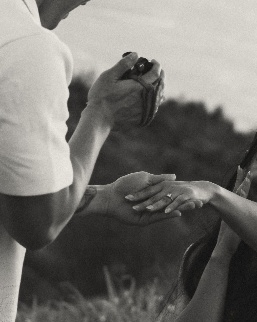 Black and white photo of a person holding a camera photographing a hand with a ring outdoors.