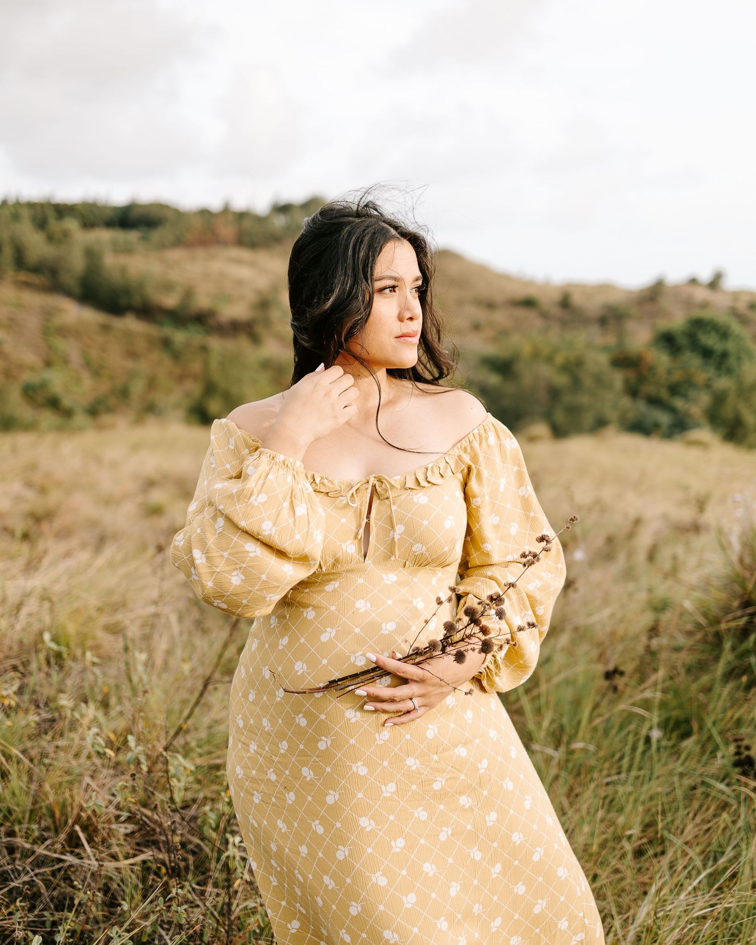 A woman in a yellow floral dress holds dried flowers in a grassy field under a cloudy sky.