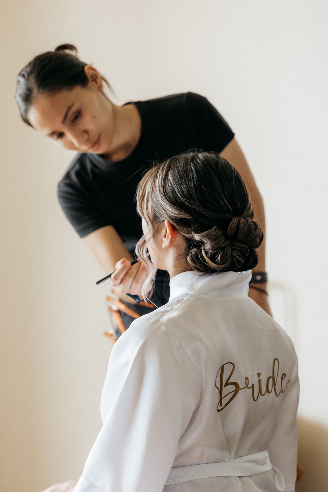 Makeup artist applying makeup to a bride wearing a white robe with gold script, bridal beauty preparation.