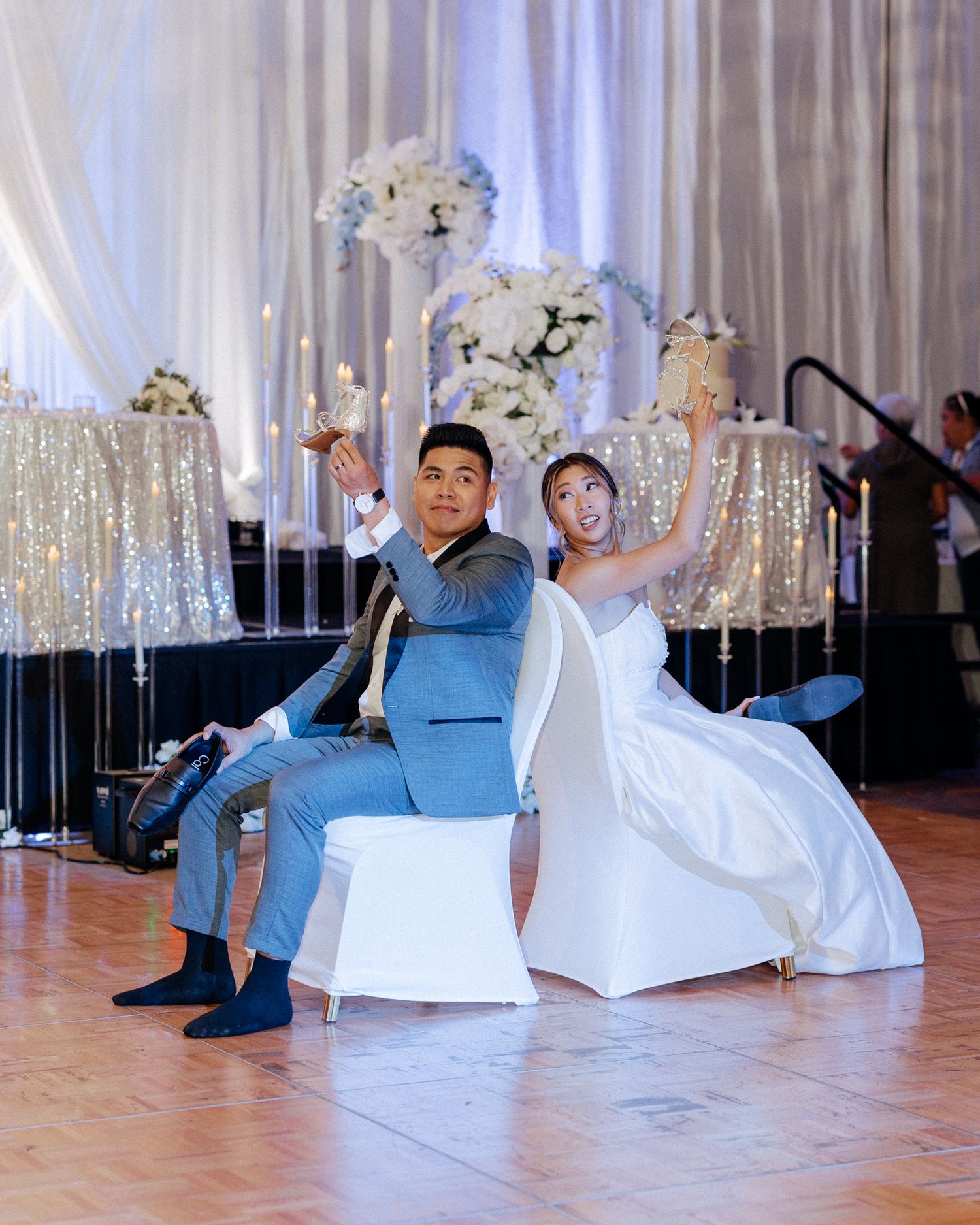A bride and groom sit back-to-back on white chairs, celebrating joyfully at their elegant wedding reception.