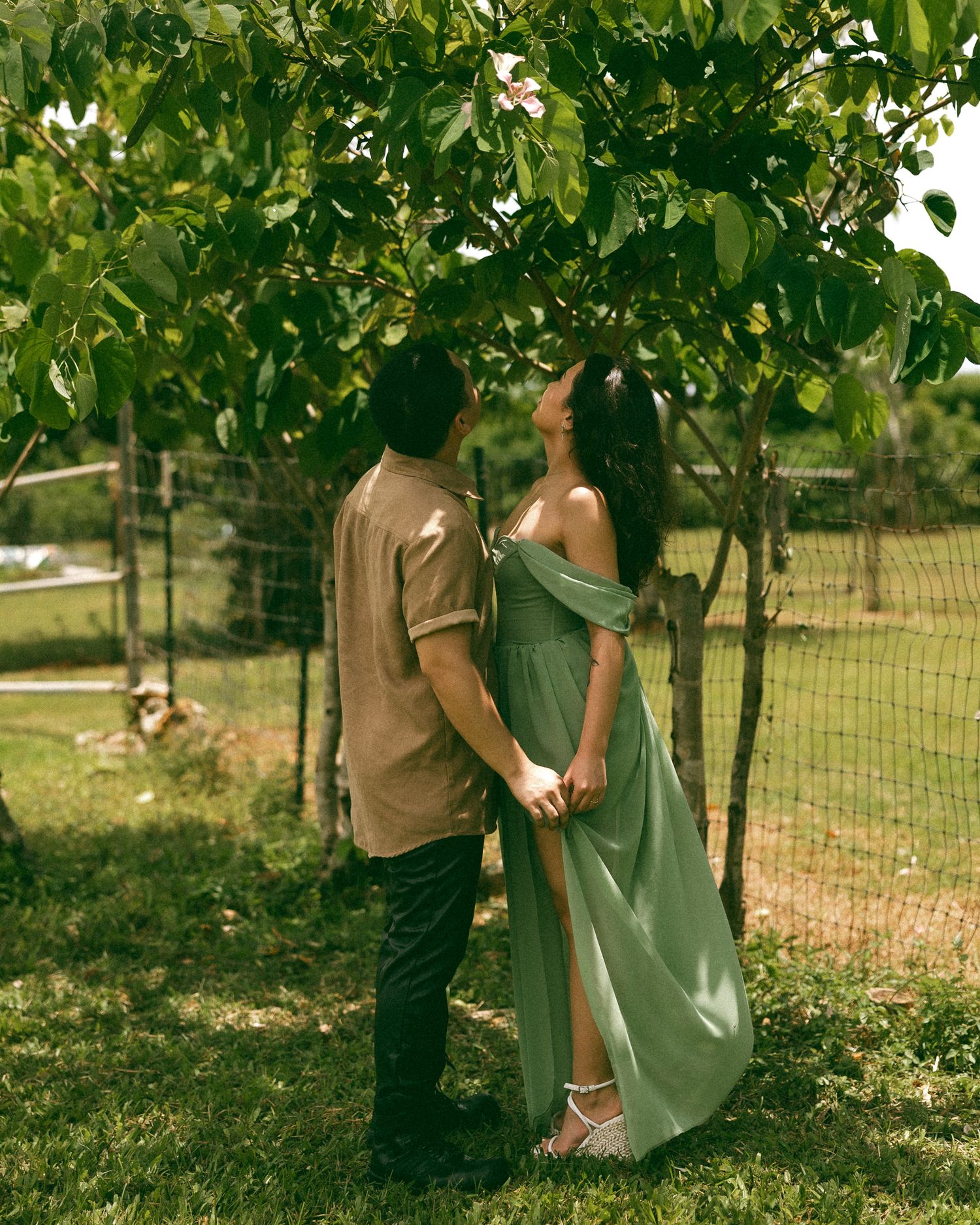 A couple holds hands and gazes at each other under a leafy tree, she wears an elegant sage green off-shoulder maxi dress.