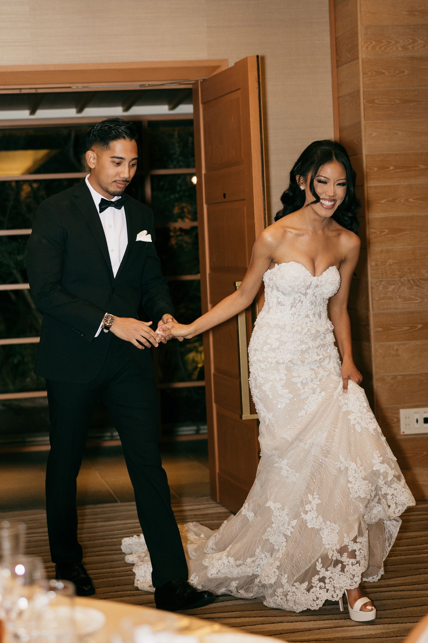 Bride in lace strapless gown and groom in black tuxedo hold hands making grand entrance at elegant wedding reception.
