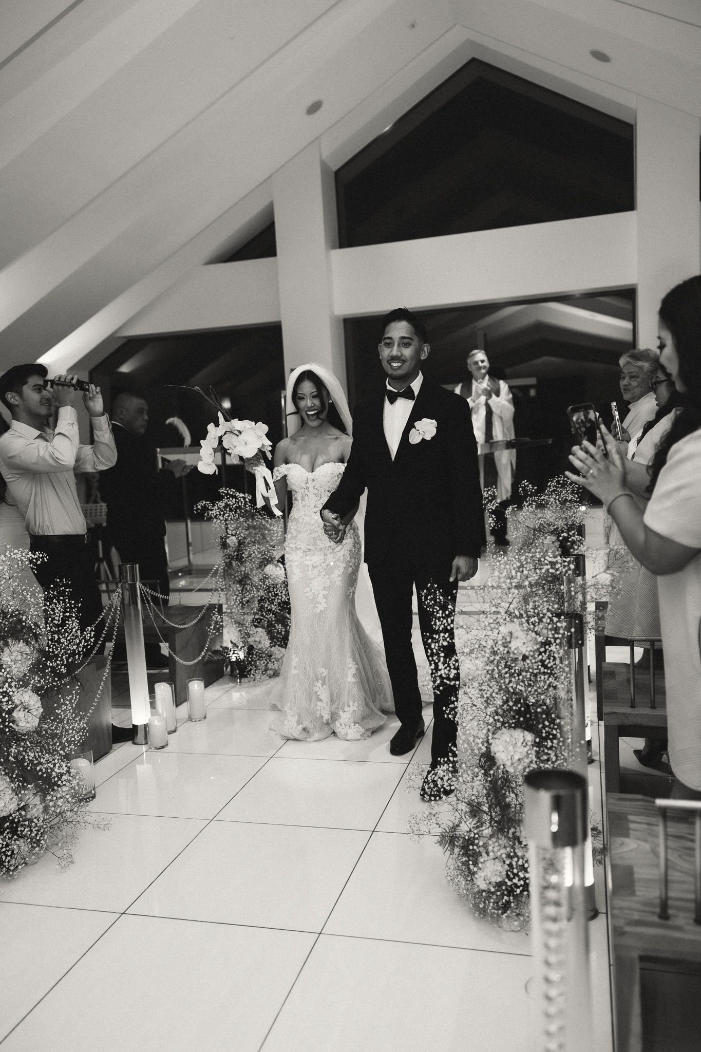 Black and white photo of a joyful couple walking down the aisle after their wedding ceremony, surrounded by floral arrangements.