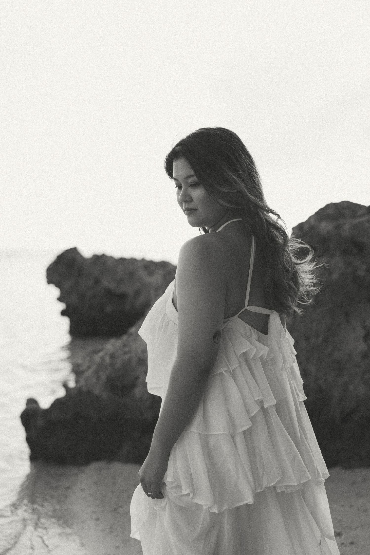 Black and white photo of a woman in a white ruffled dress standing on a rocky beach, hair blowing in the wind.