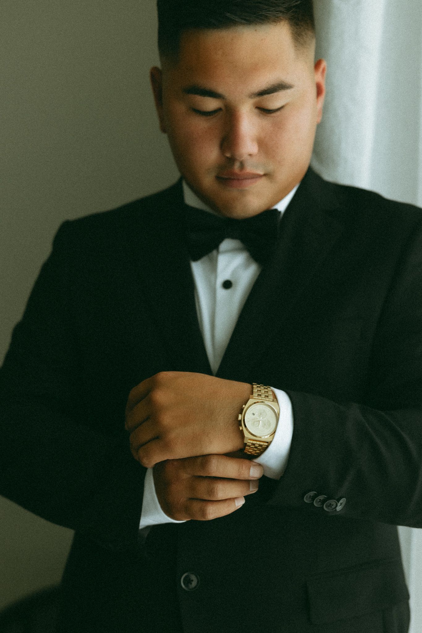 A groom in a black tuxedo with bow tie adjusts his gold watch cufflinks while preparing for his wedding.