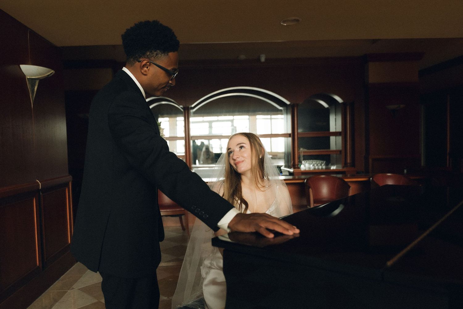 Groom in black suit plays piano with bride in wedding dress seated beside him in elegant venue.
