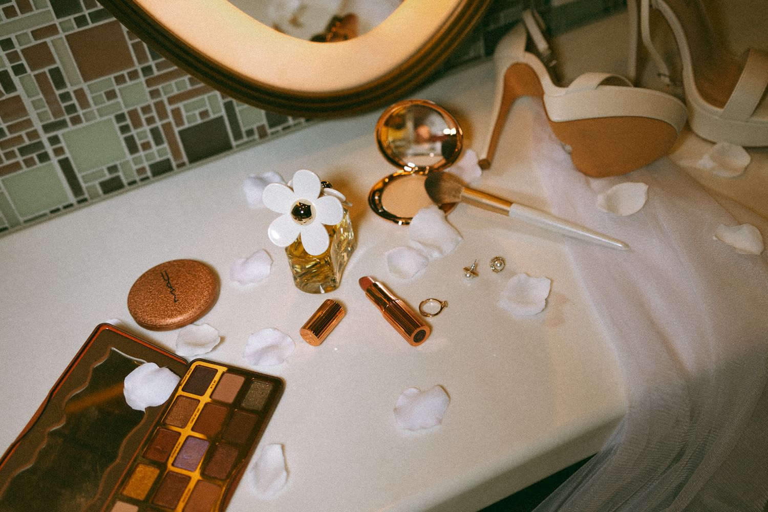Luxury makeup and beauty products with gold accents arranged on a white bathroom sink with flower petals.