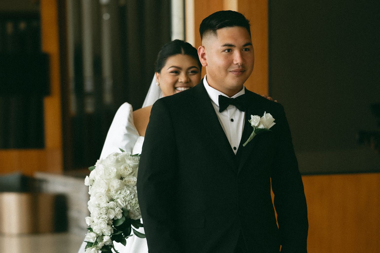 Bride and groom in elegant wedding attire, groom in black tuxedo with white boutonniere, bride holding white floral bouquet.