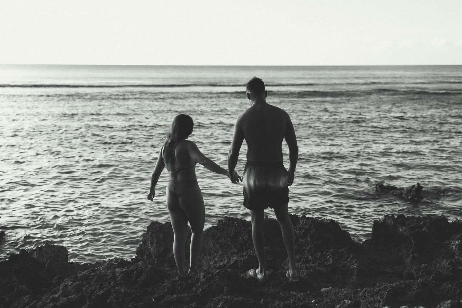 Black and white photo of a couple holding hands on rocky shore, gazing at the ocean horizon together.