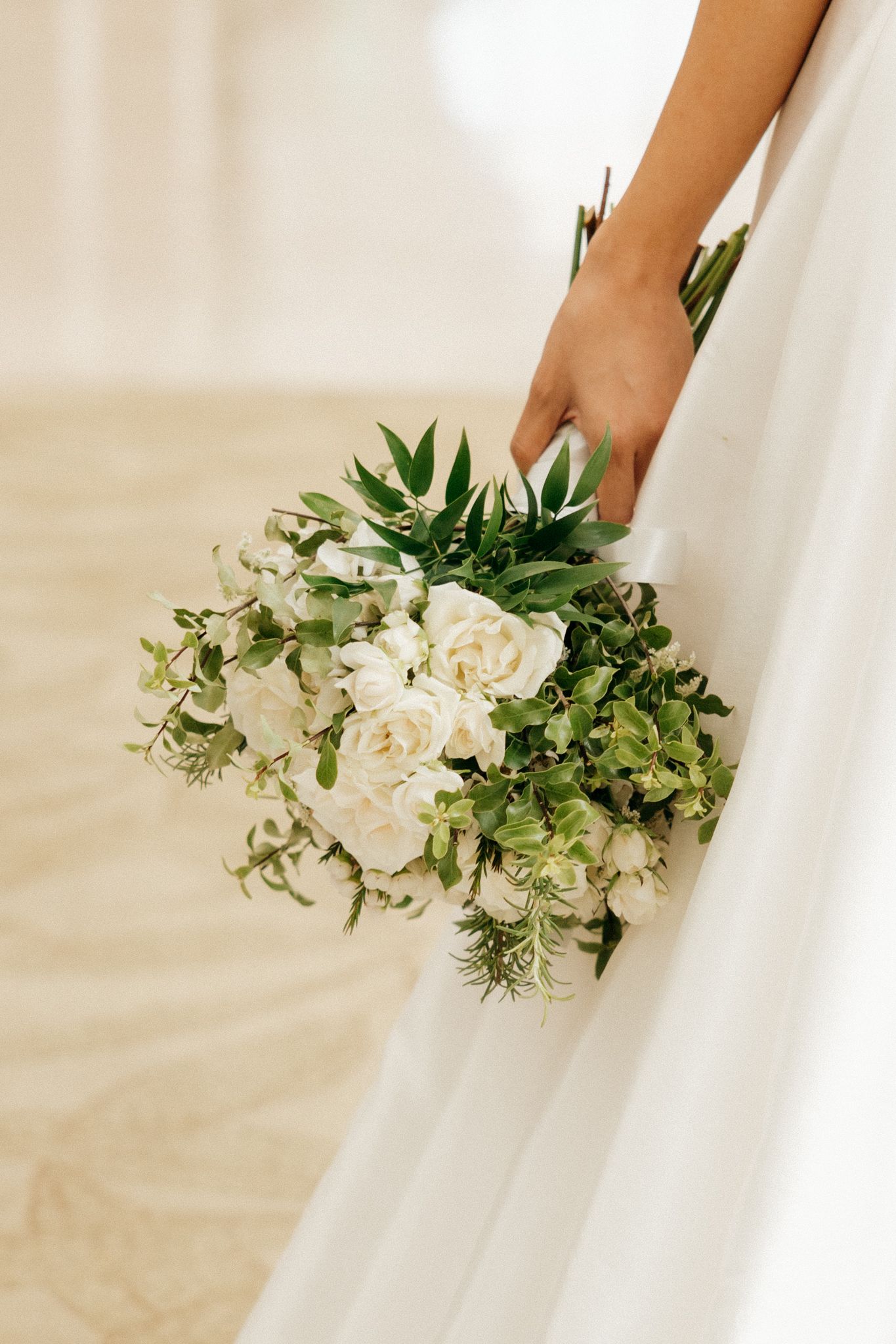 Bride holding a lush bridal bouquet of white roses and green foliage against a flowing white wedding dress.