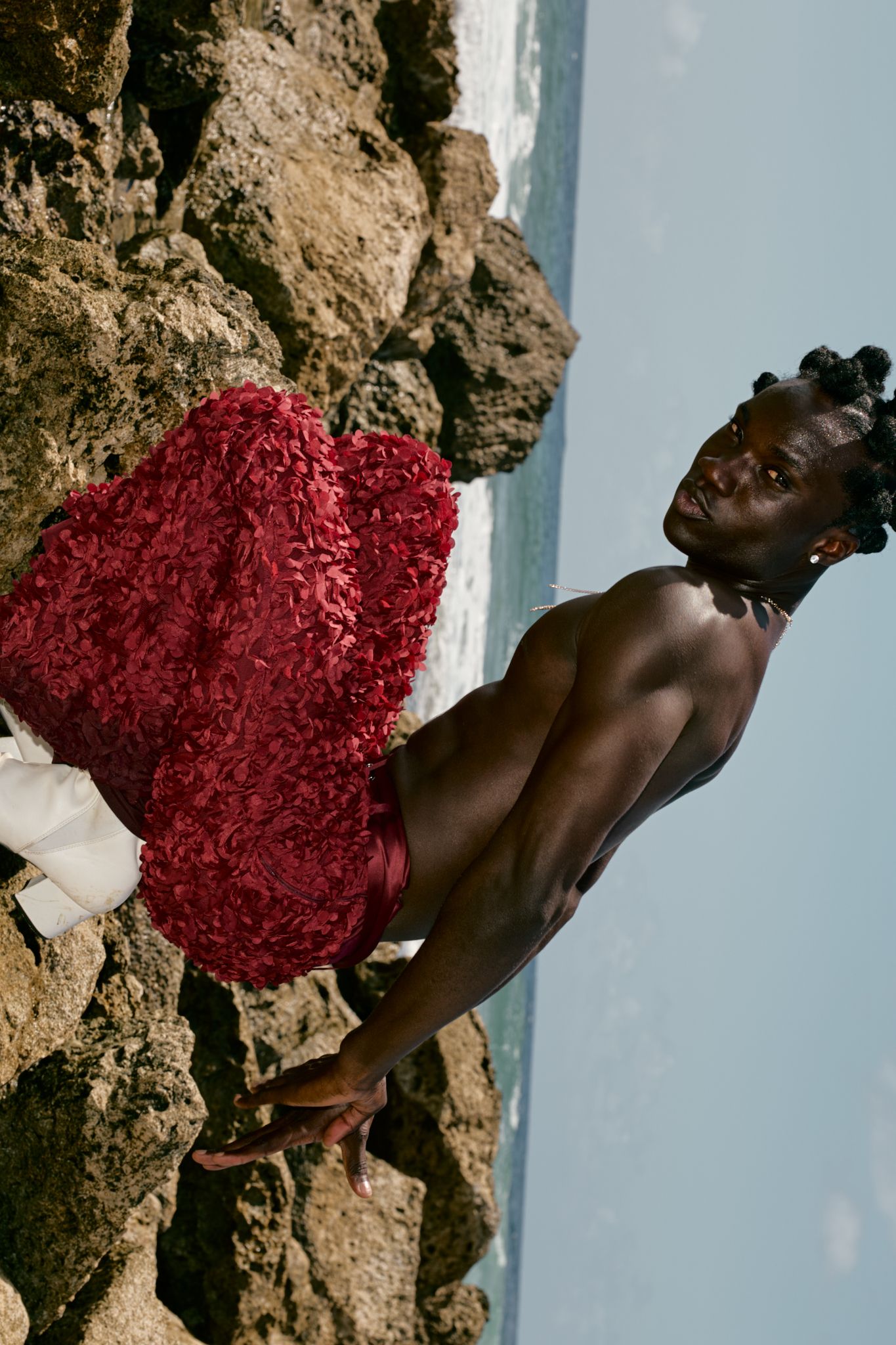 Shirtless Black man in red ruffled shorts poses against rocky coastal cliffs with ocean in background.