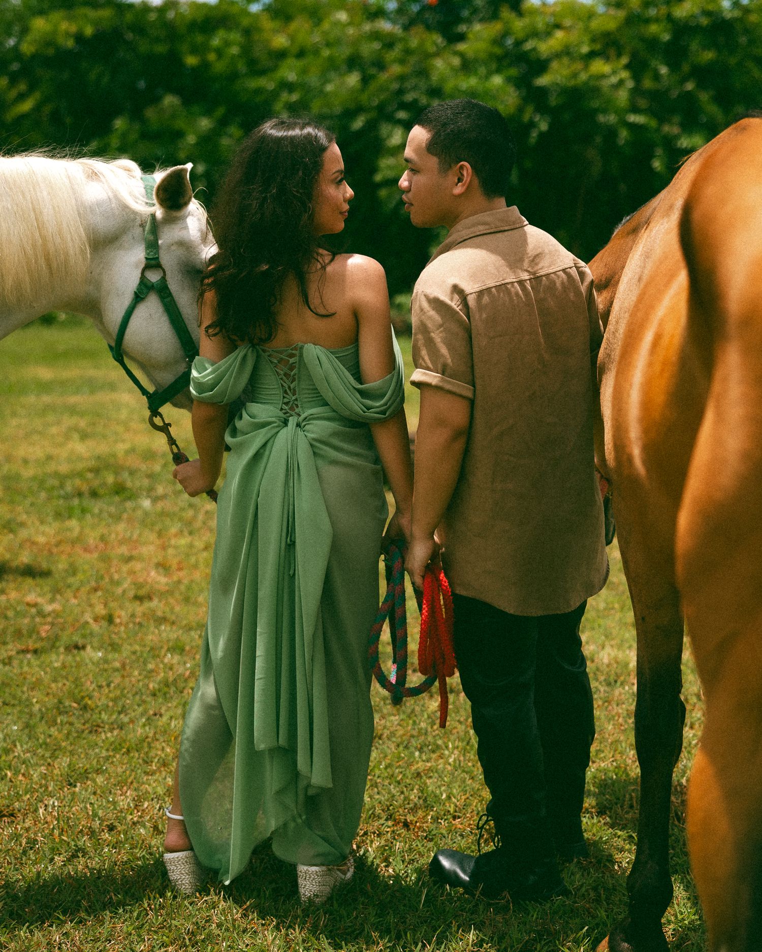 A couple in elegant attire shares a romantic moment between a white and brown horse on a lush green field.