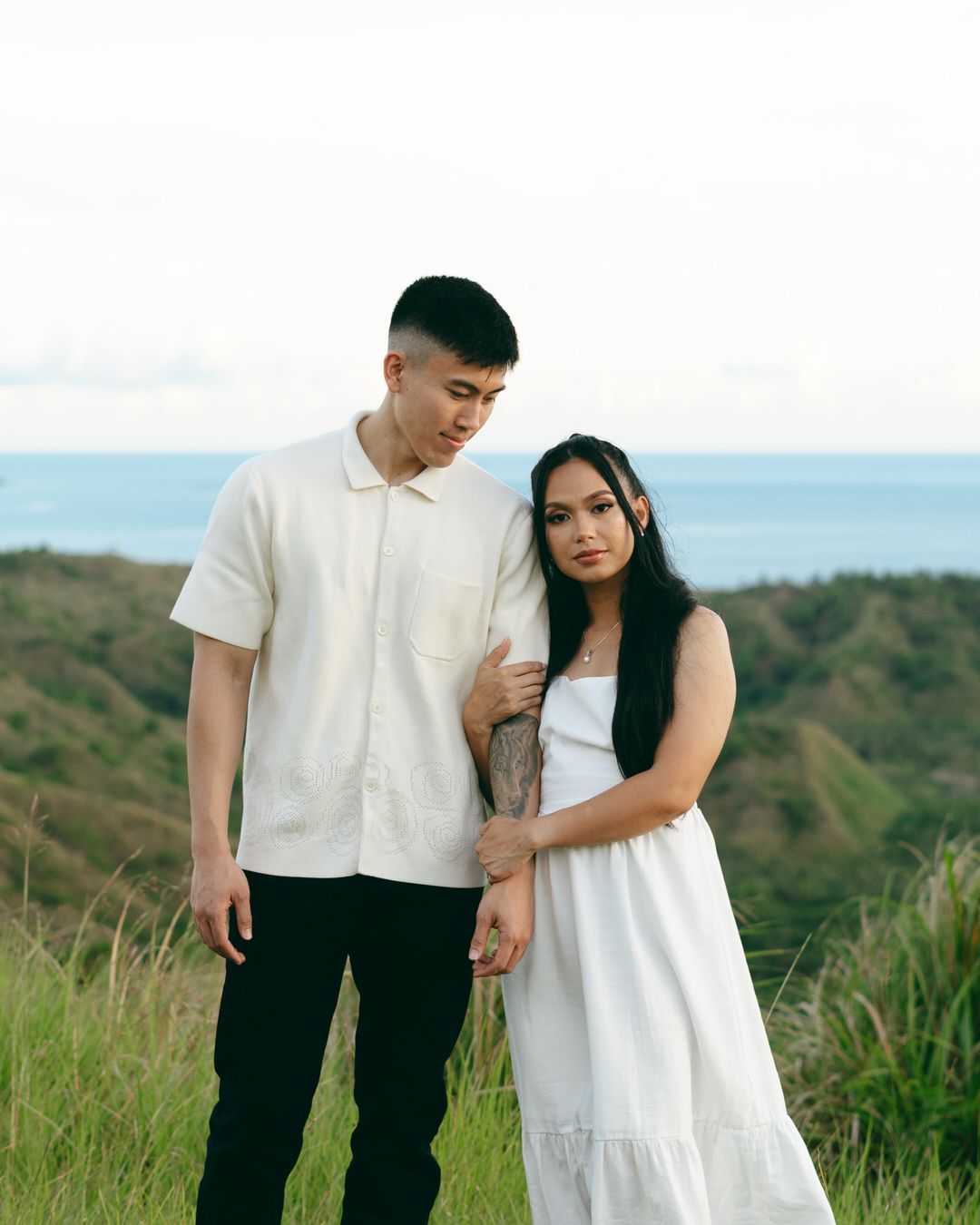 A young couple in white outfits poses on a grassy hillside with ocean views in the background.