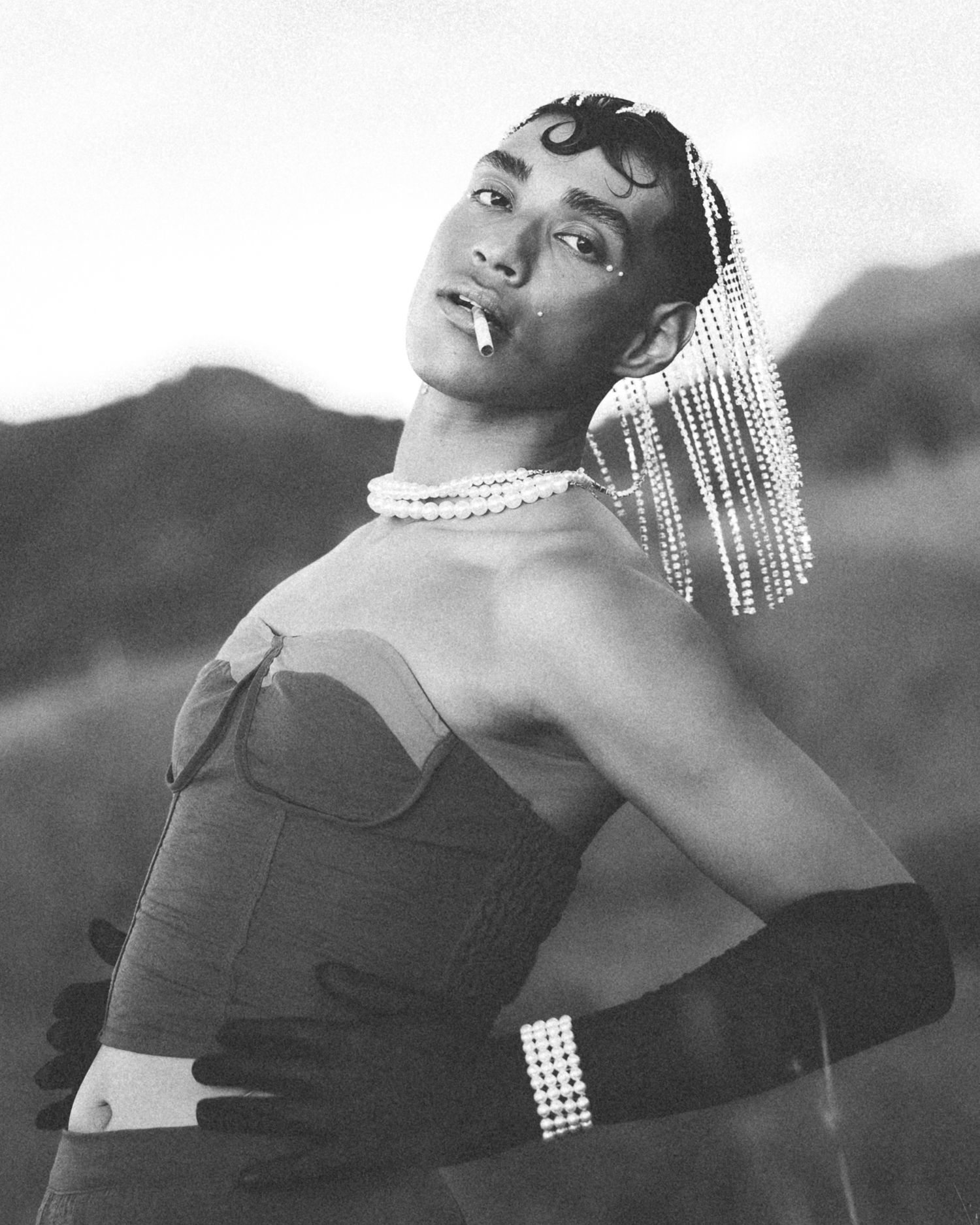 Black and white photo of a glamorous person in strapless top, pearl jewelry, and long gloves posing outdoors.