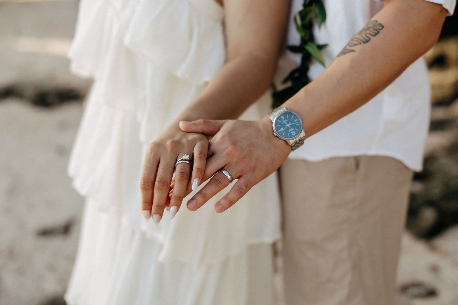 Newlyweds displaying wedding rings and a blue watch on a beach, wearing white dress and khaki pants.
