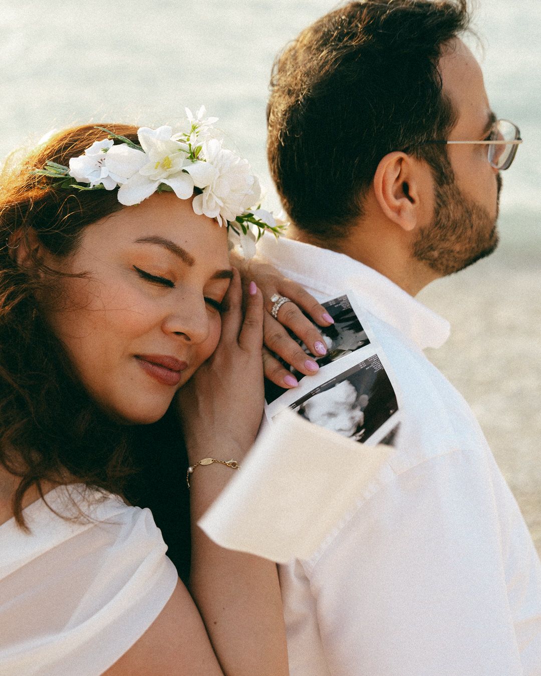 Couple sharing a tender moment, woman with floral crown resting her head on man's shoulder holding an ultrasound photo.