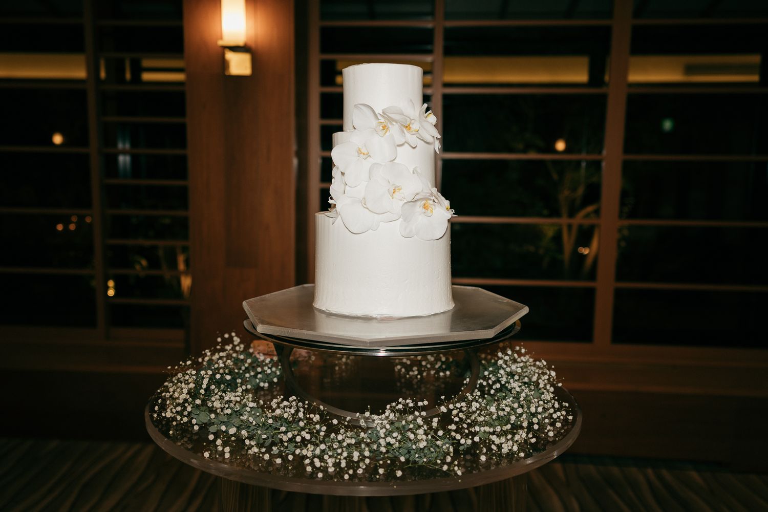 Elegant three-tier white wedding cake adorned with florals, displayed on a mirrored stand surrounded by baby's breath.