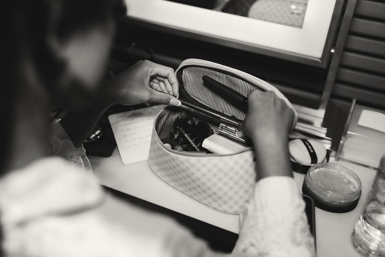 Black and white photo of a person looking into an open makeup bag on a vanity table.