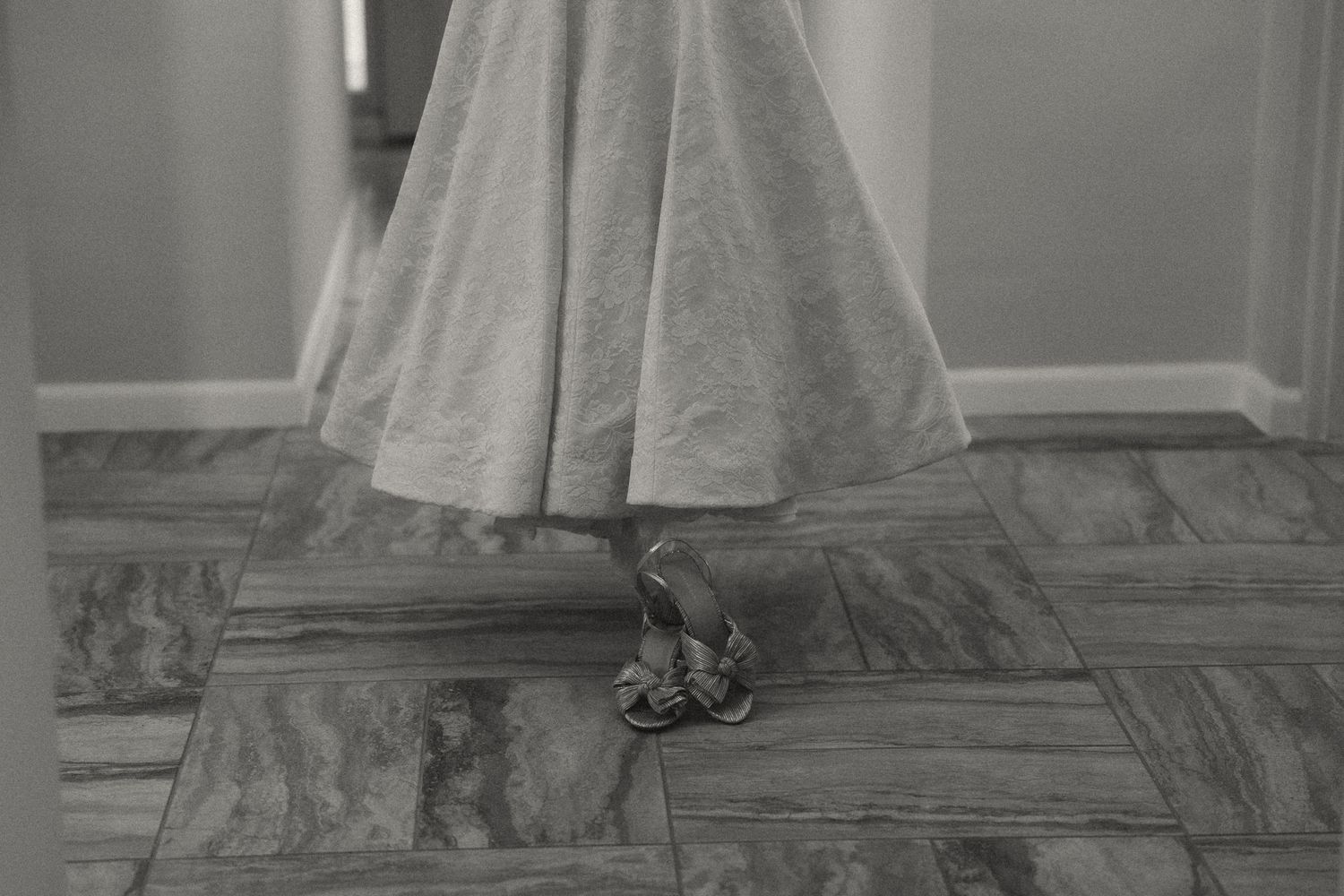 Black and white photo of a woman in a full-length skirt and heels standing on a tiled floor near a mirror.