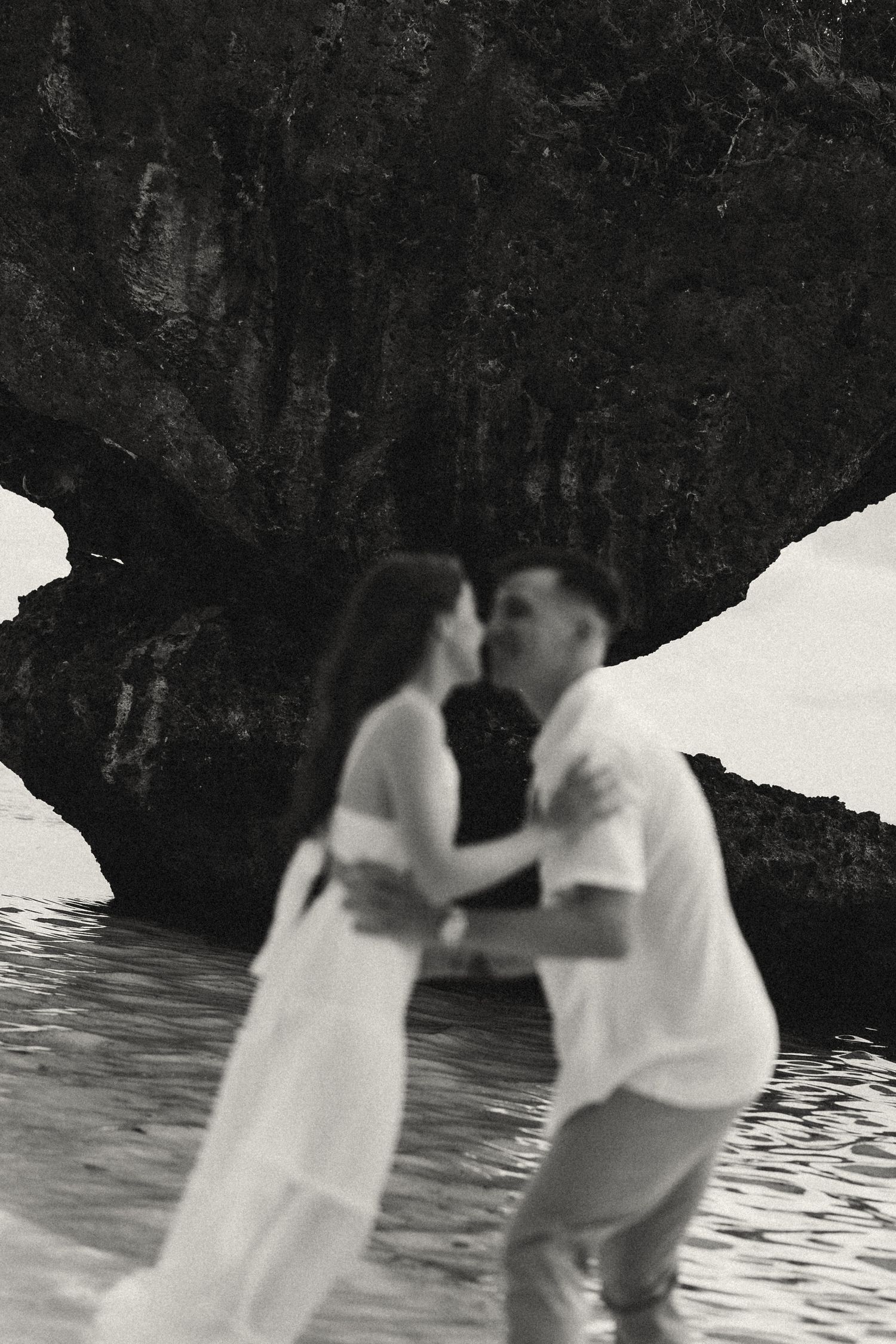 Black and white photo of a couple embracing and kissing near rocky cliffs by the ocean water.