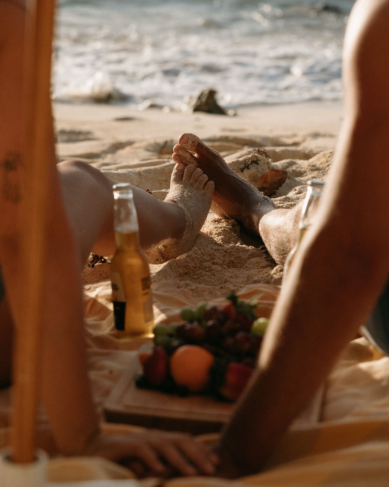 People relaxing on a sandy beach with beer bottles and a fruit platter on a blanket near the ocean.