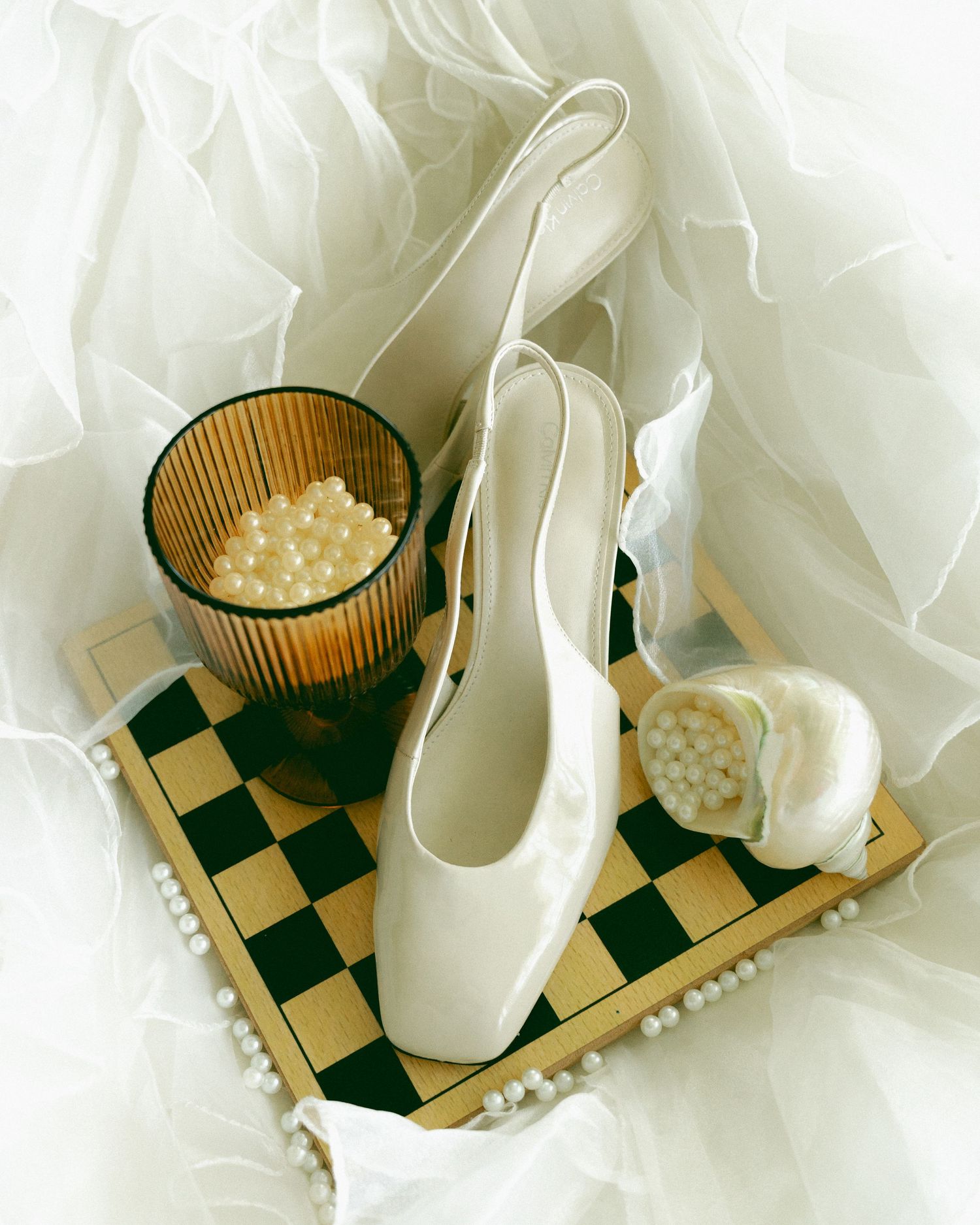 White satin bridal heels on a chess board with pearls and a candle, surrounded by tulle fabric.