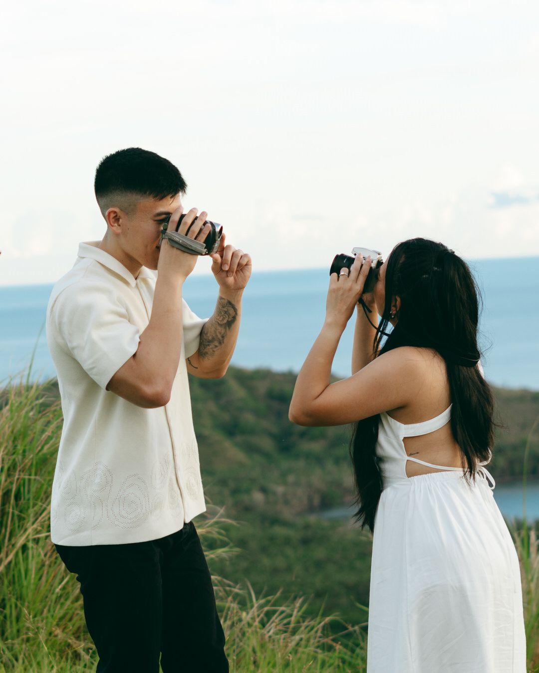 A couple photographs each other with cameras on a scenic hilltop overlooking the ocean, dressed in white outfits.