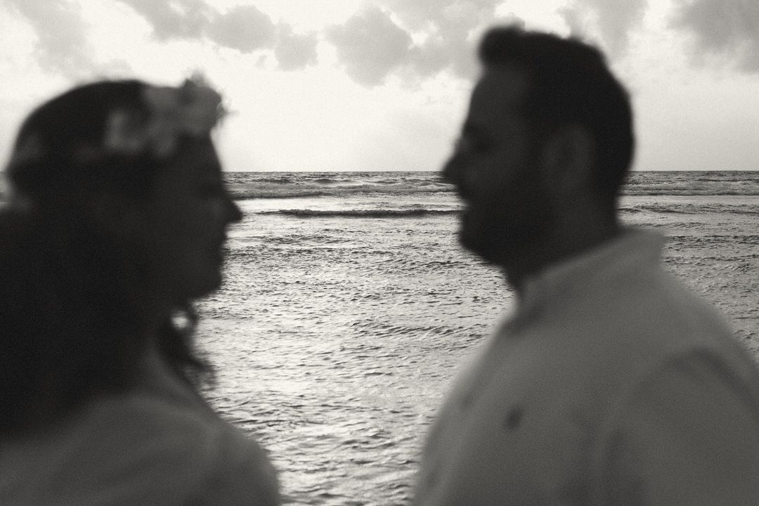 Black and white photo of a couple facing each other on a beach at sunset, woman wearing a flower crown.