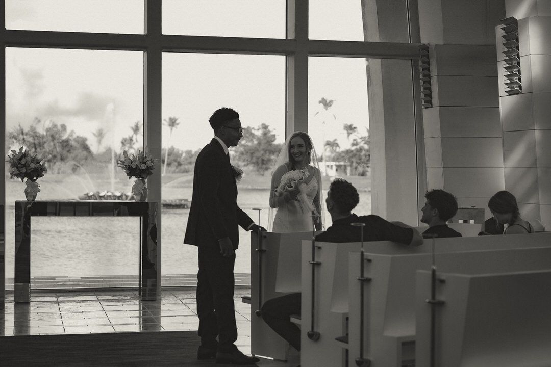Black and white wedding ceremony photo of bride and groom holding hands at altar with guests seated nearby.