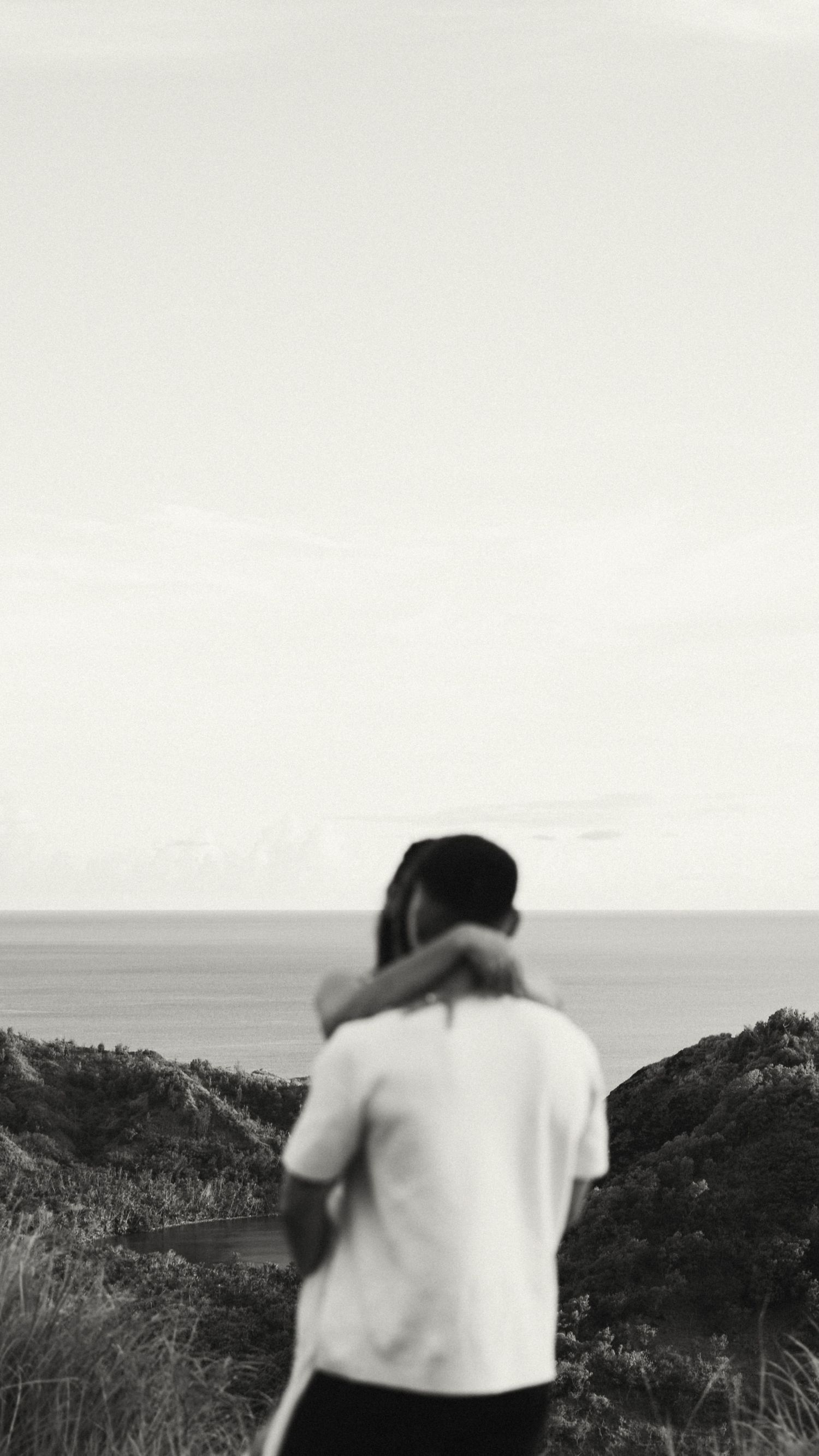 Black and white photo of a couple embracing on a coastal hilltop with ocean horizon in background.