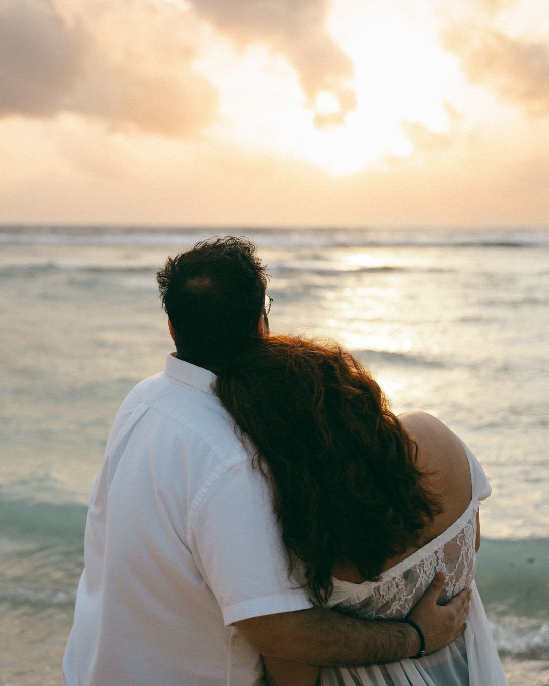 Couple embracing on a tropical beach at sunset, watching golden waves under a dramatic cloudy sky.