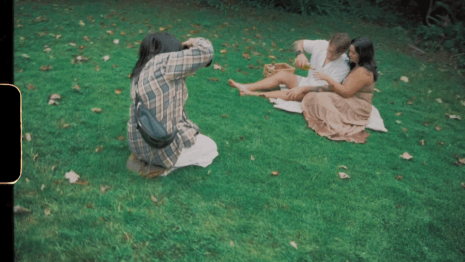 Person photographing two women relaxing on green grass in a sunny backyard garden setting.