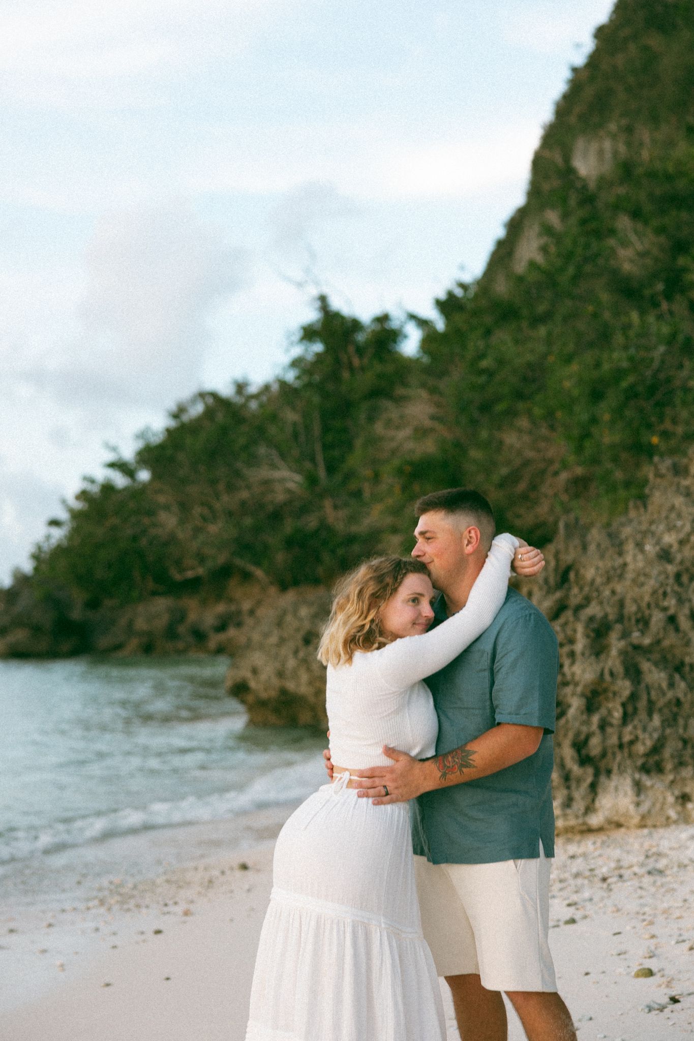 A couple embraces tenderly on a tropical beach, with rocky cliffs and turquoise water in the background.