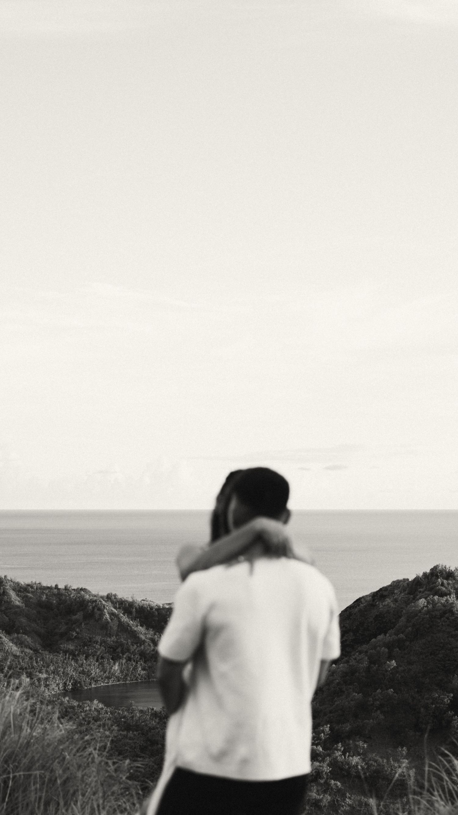 Black and white photo of a couple embracing on a coastal hillside with ocean views in the background.