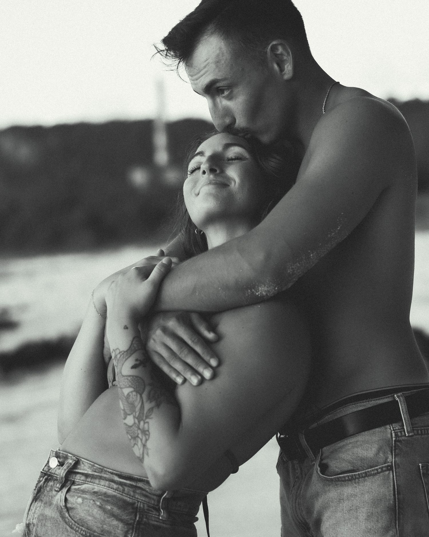 Black and white photo of a shirtless couple embracing outdoors, man kissing womans head near water.