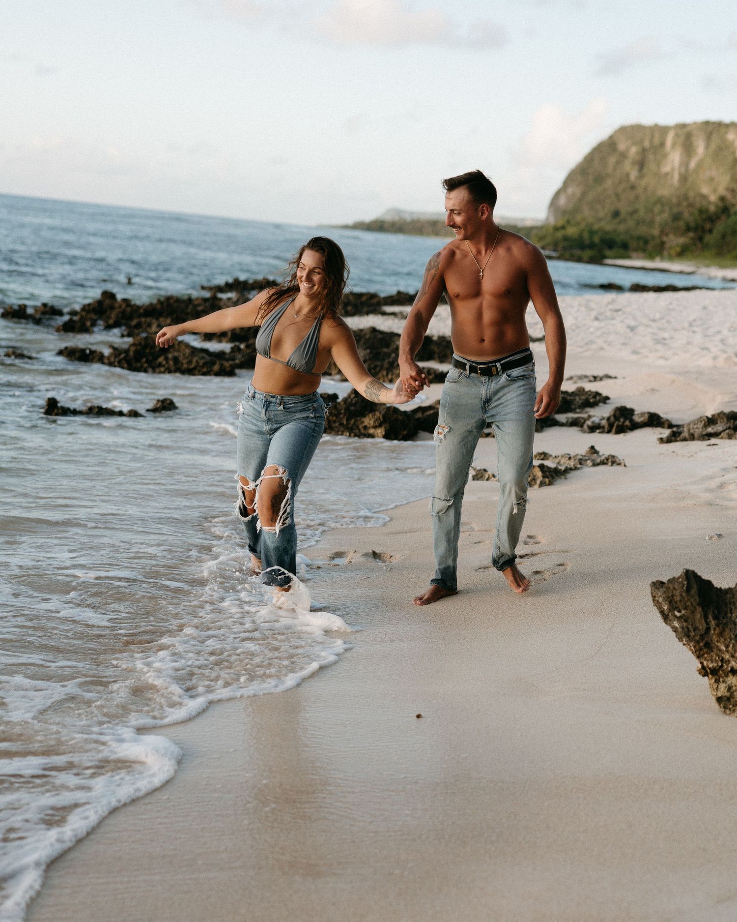 A couple holding hands and laughing while walking along a sandy beach with ocean waves at their feet.