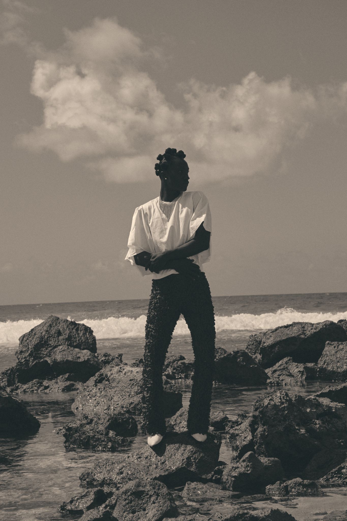 A person stands on rocky shoreline in sepia-toned photo, wearing white tee and dark jeans, with ocean waves and cloudy sky.