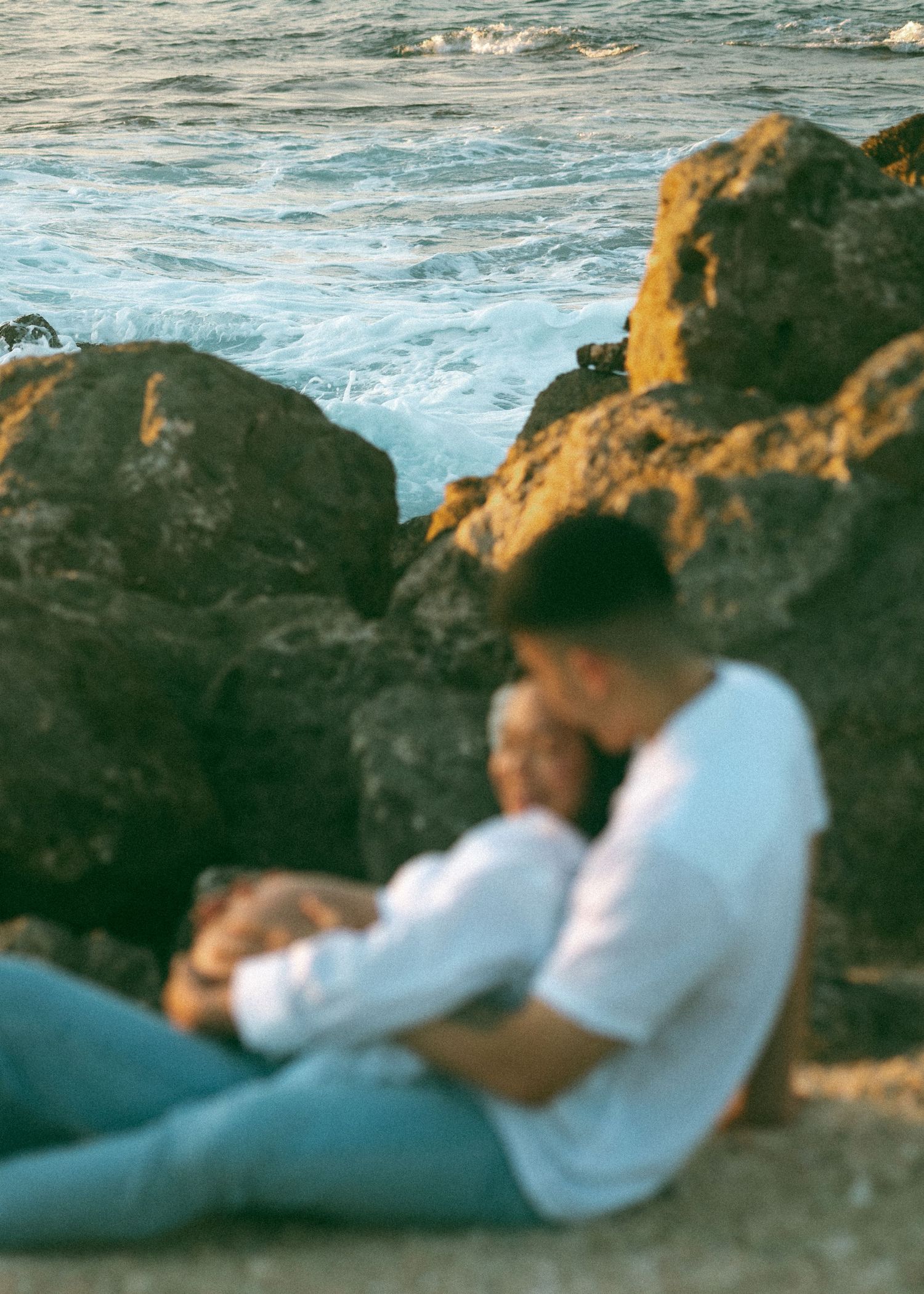 Couple sitting together on rocky beach shore at golden hour, sharing a tender embrace by the ocean waves.