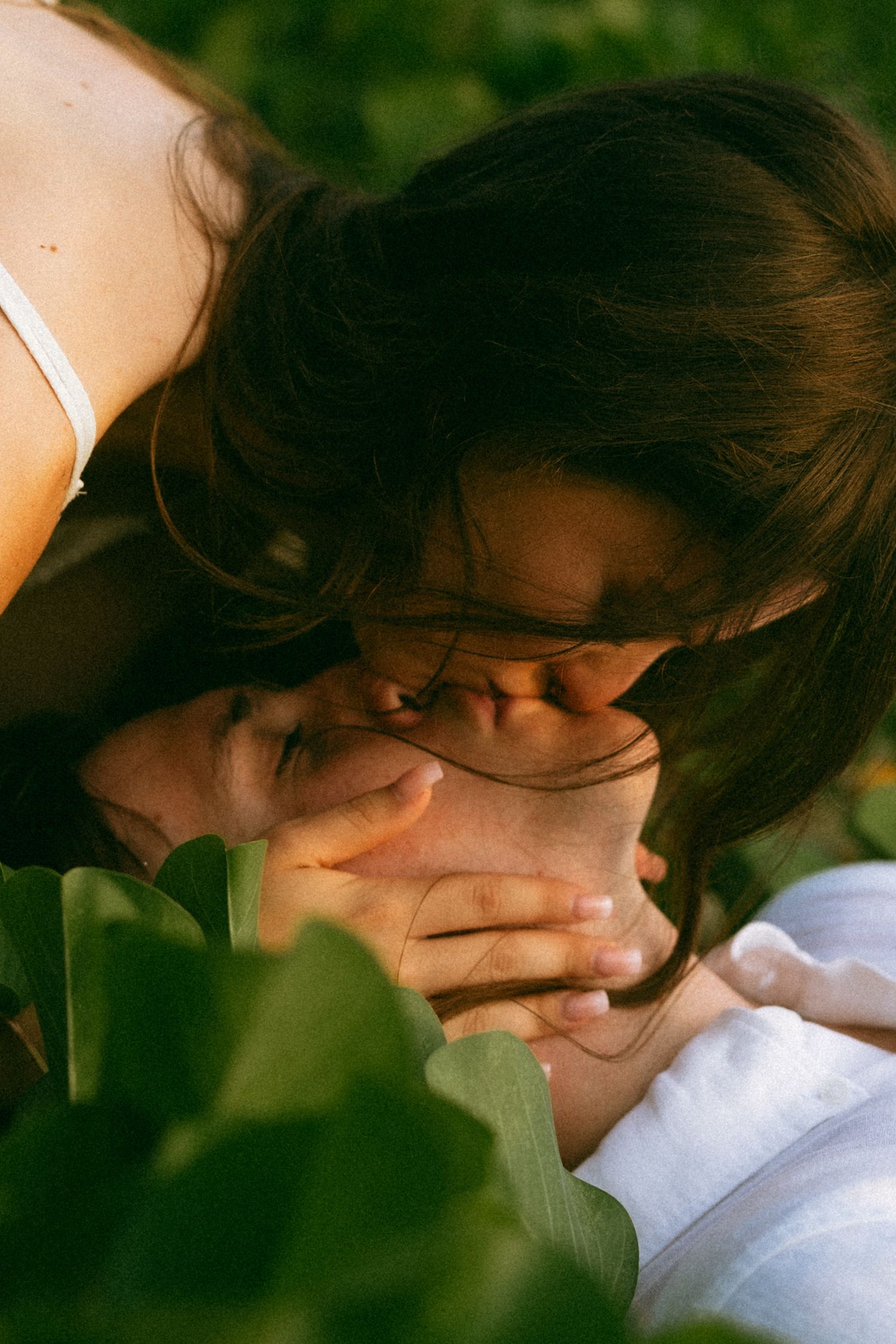 Two people sharing a tender kiss outdoors, surrounded by lush green foliage in warm natural light.