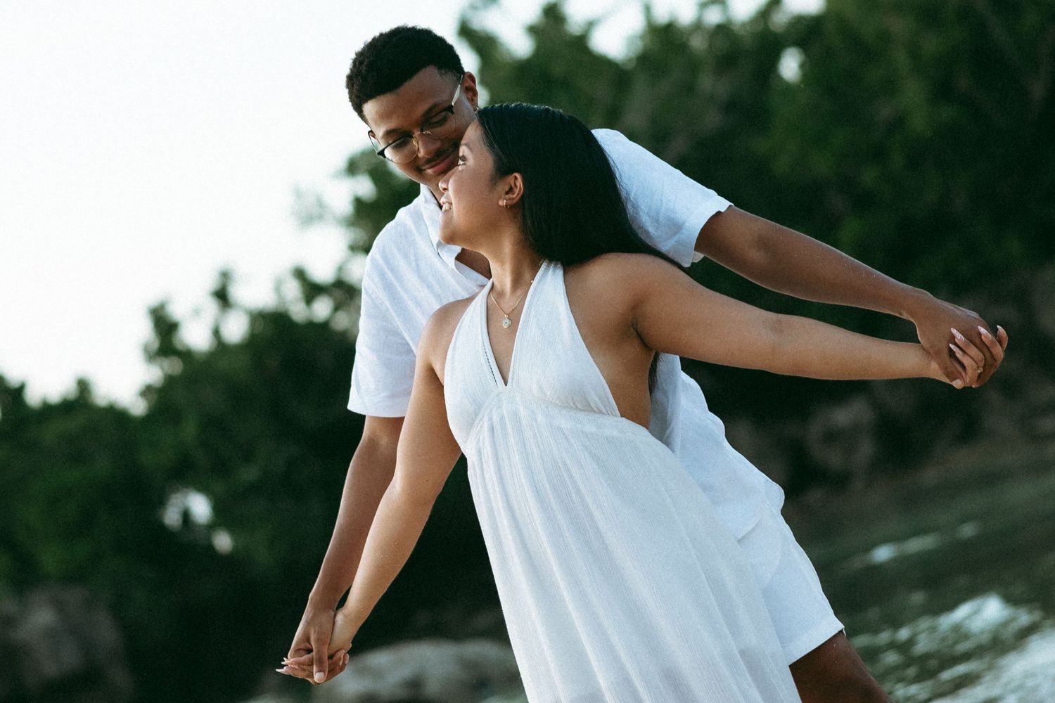 A couple in white outfits dancing romantically on a tropical beach, smiling and holding hands.