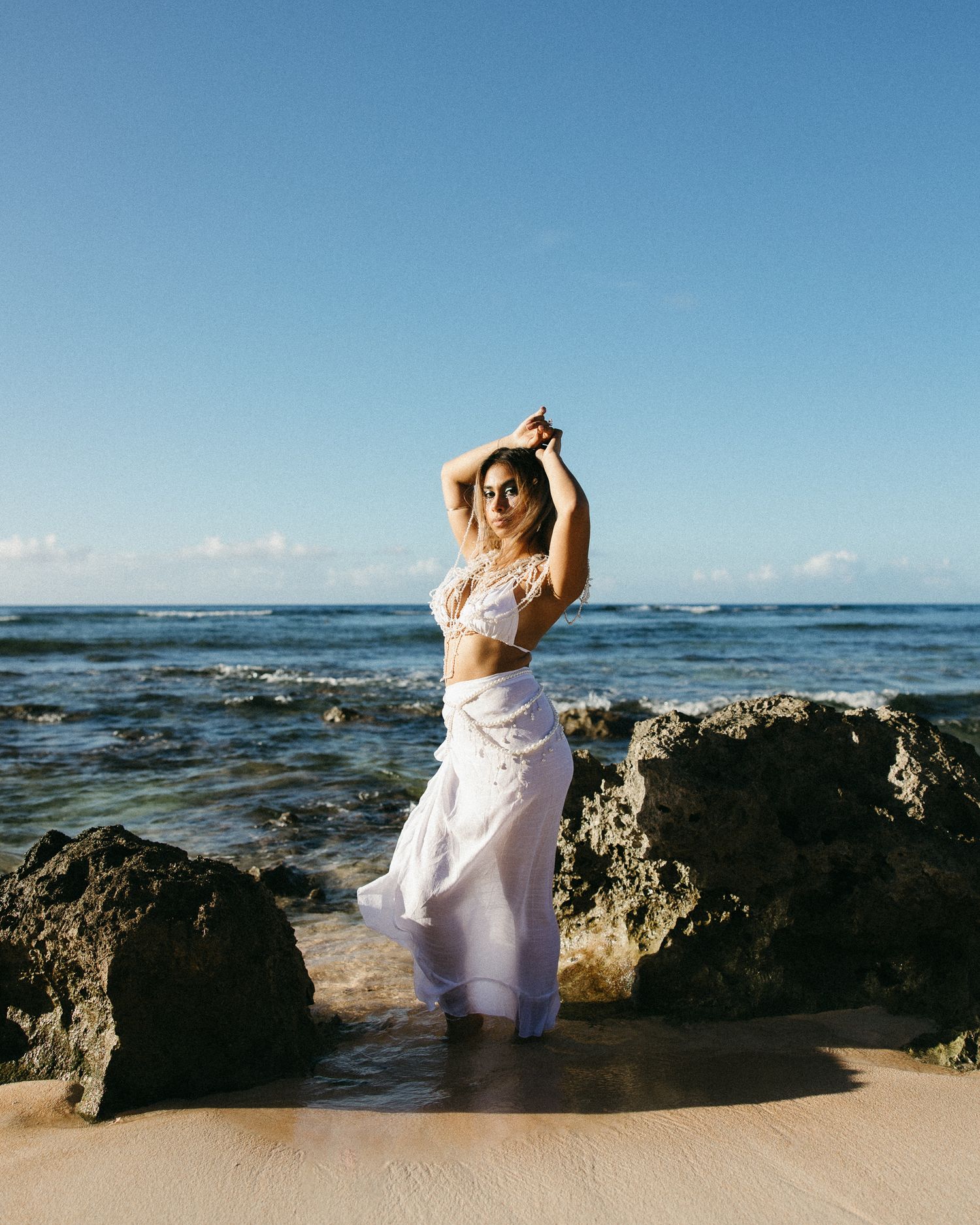 Woman in white outfit posing on rocky beach with ocean waves and blue sky backdrop.