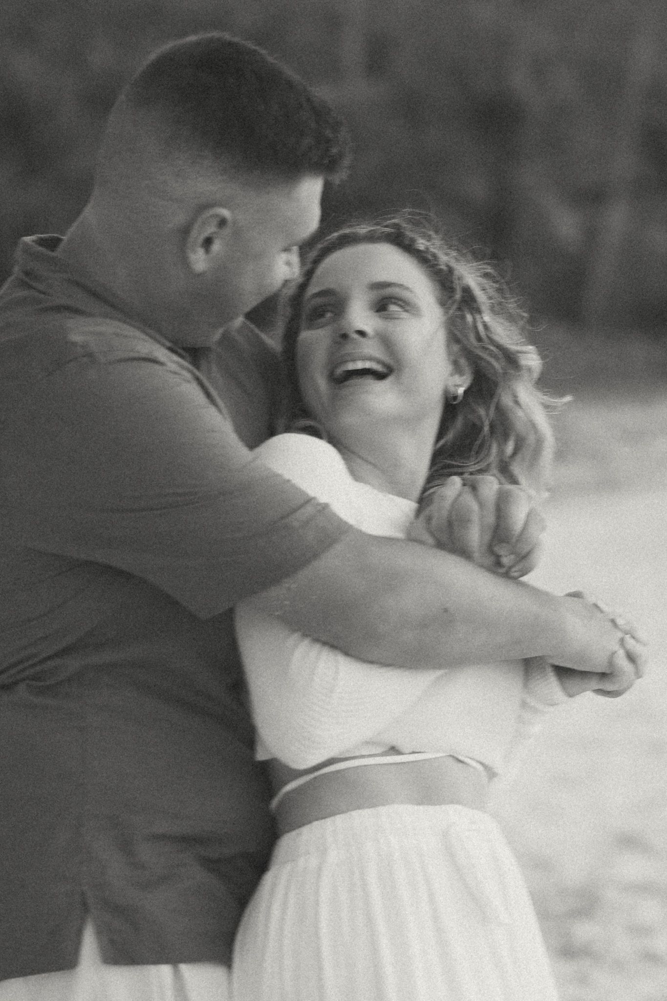 Black and white photo of a couple embracing and laughing outdoors, woman in white dress, man in casual shirt.