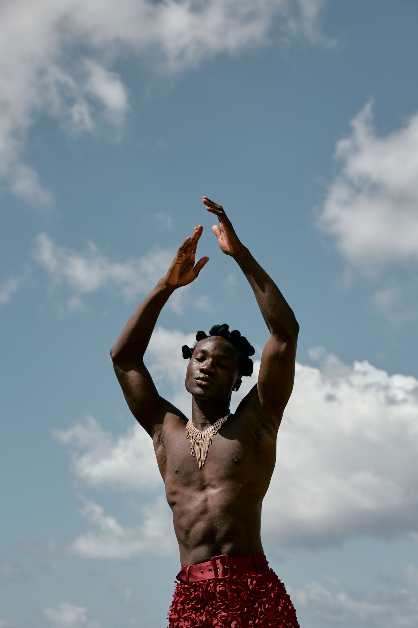 Shirtless Black male dancer raises arms gracefully against cloudy blue sky, wearing red sequin pants and gold necklace.