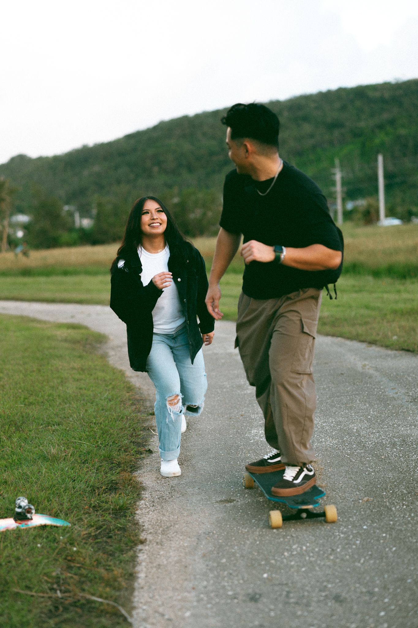 Young couple enjoying outdoors, woman running alongside man skateboarding on a path with green hills in background.