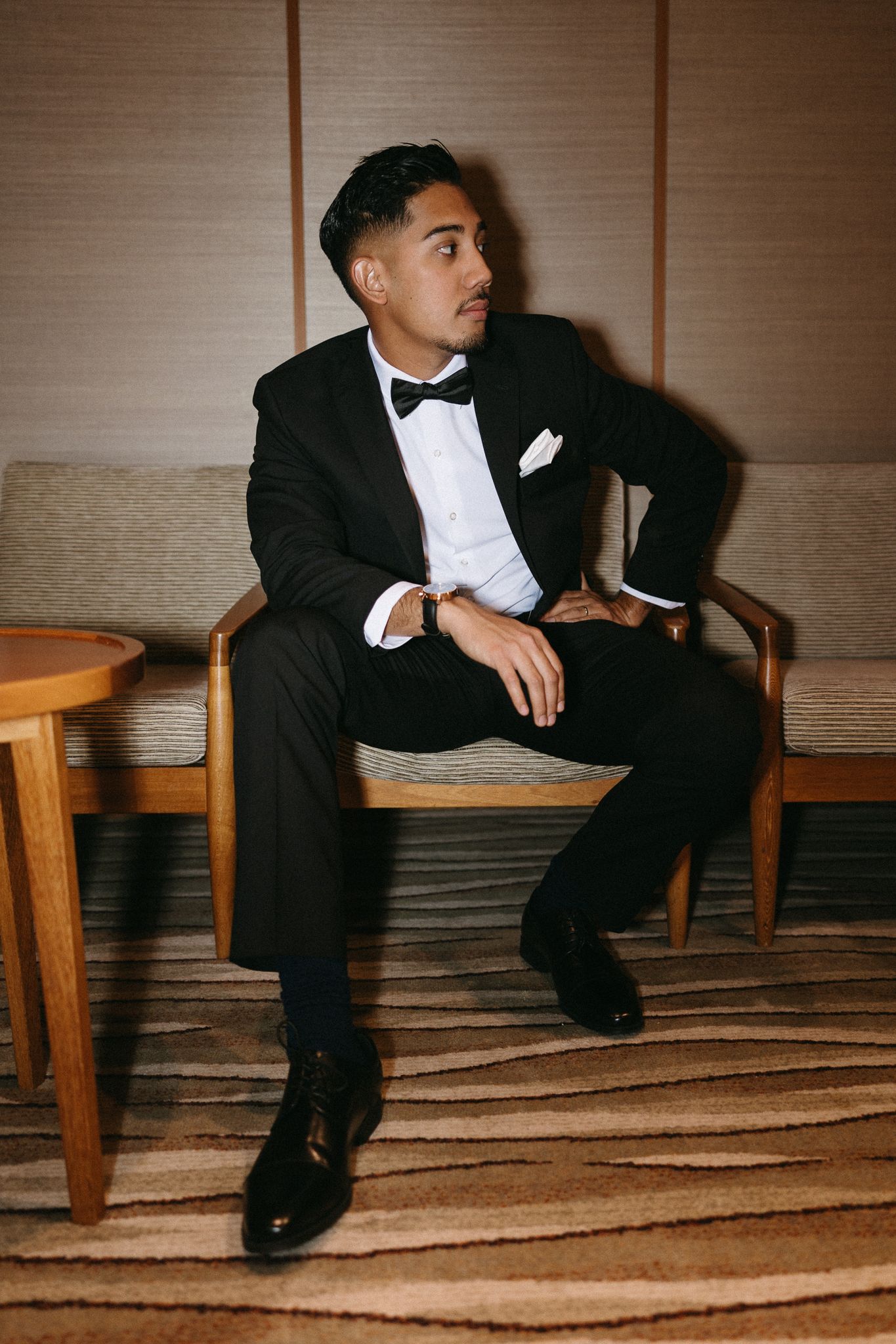 Elegant man in black tuxedo with bow tie seated in a wooden chair, posing confidently in a formal setting.