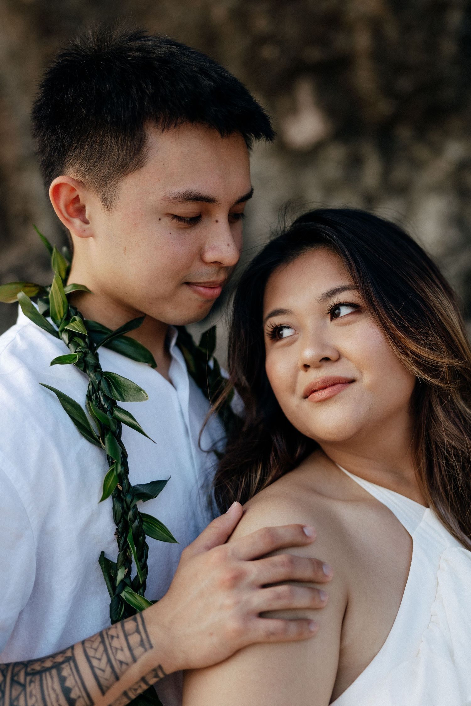 A couple in white attire share a tender moment, the man wearing a green lei and showing a tattoo on his arm.