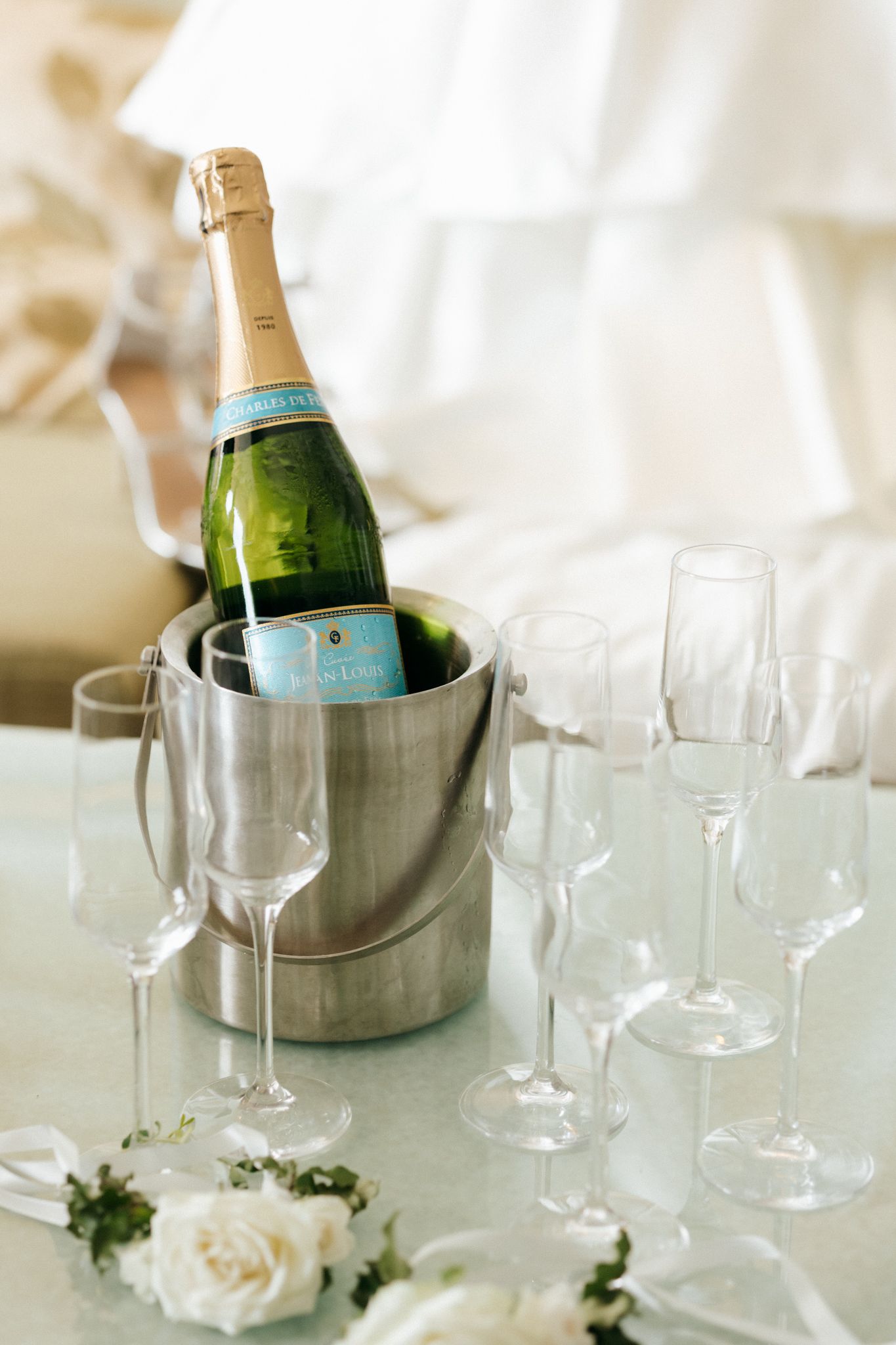 Champagne bottle chilling in silver ice bucket surrounded by empty flute glasses and white roses on a table.