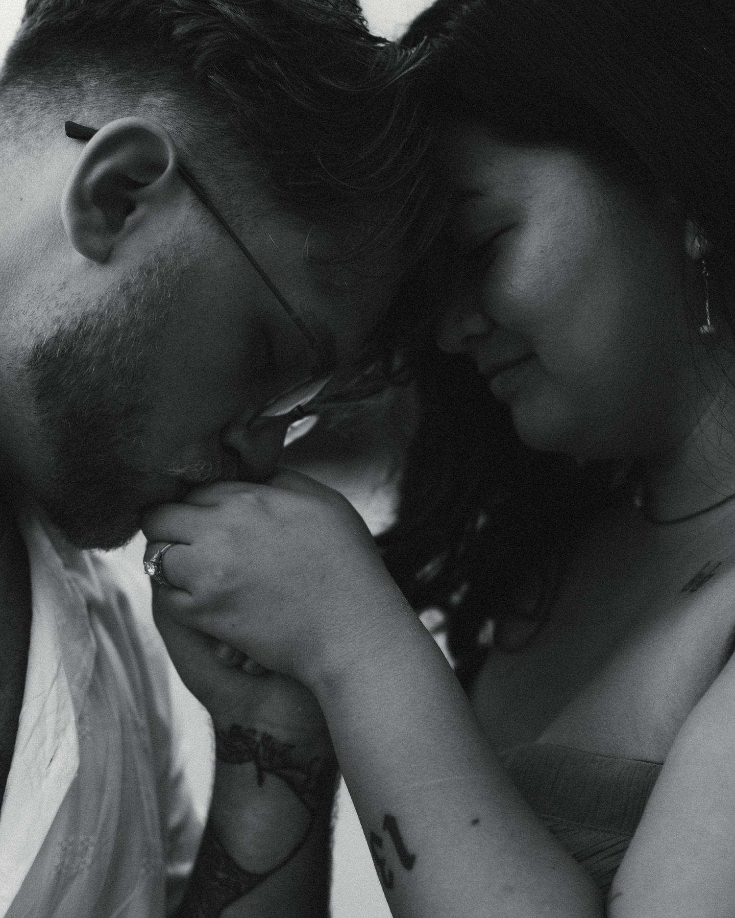 Black and white photo of a man kissing a woman's hand tenderly, both with tattoos, sharing a romantic intimate moment.