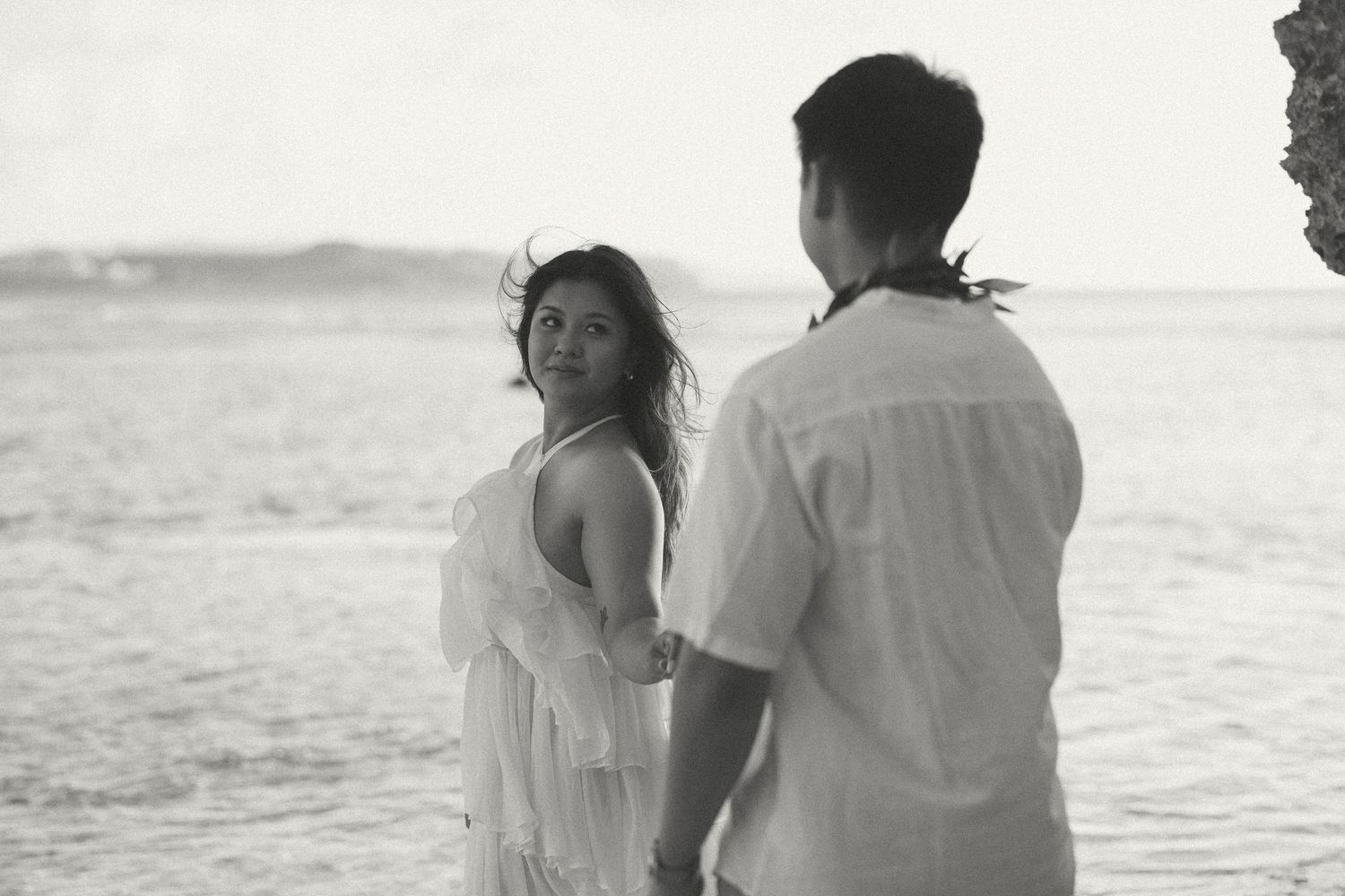 A couple holds hands on a beach in a black and white photo, woman in white dress looks back romantically.