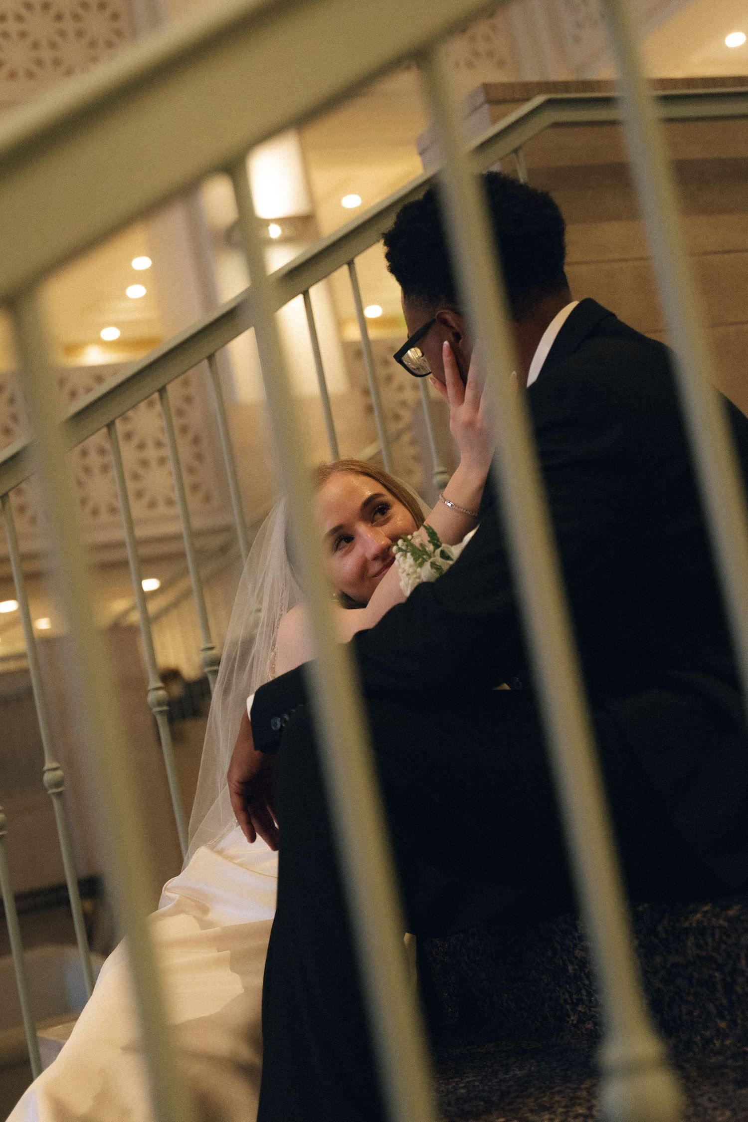 Bride in white veil and groom in black suit posing on elegant staircase at wedding venue.